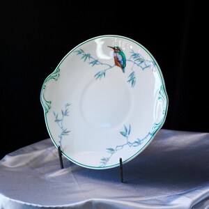 May include: White ceramic plate with a green rim and decorative handles. The plate features a painted design of a colorful bird perched on a branch with blue leaves. The plate is displayed on a black metal stand.