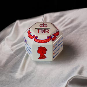 Op de afbeelding: Een achthoekige witte keramische spaarpot met rode en blauwe tekst. De tekst bevat "ER", "Elizabeth II" en "God save the Queen!". De doos heeft ook een silhouet.