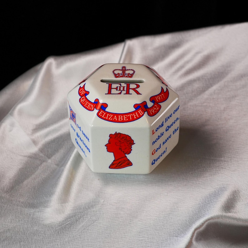 Op de afbeelding: Een achthoekige witte keramische spaarpot met rode en blauwe tekst. De tekst bevat "ER", "Elizabeth II" en "God save the Queen!". De doos heeft ook een silhouet.