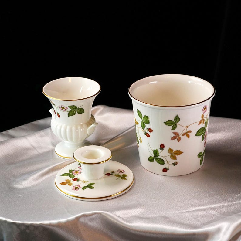 Op de afbeelding: Een set witte keramische decoratieve items met een aardbeienmotief. De set bevat een kleine vaas, een kandelaar en een cilindrische container, allemaal met gouden randen. Het aardbeienontwerp heeft groene bladeren en rode bessen.