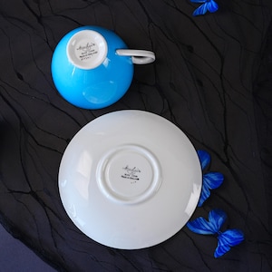 Op de afbeelding: Een felblauwe theekop van bone china en een bijpassende witte schotel, beide voorzien van het Mayfair-merkstempel en de tekst Made in England aan de onderzijde. De theekop heeft een gebogen handvat en de schotel heeft een ronde inkeping in het midden.