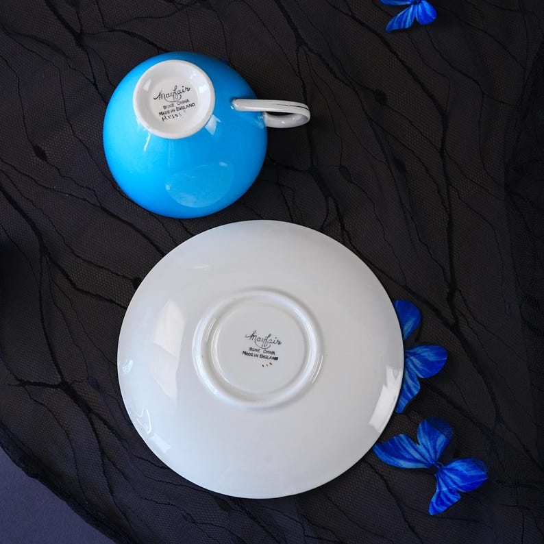 Op de afbeelding: Een felblauwe theekop van bone china en een bijpassende witte schotel, beide voorzien van het Mayfair-merkstempel en de tekst Made in England aan de onderzijde. De theekop heeft een gebogen handvat en de schotel heeft een ronde inkeping in het midden.