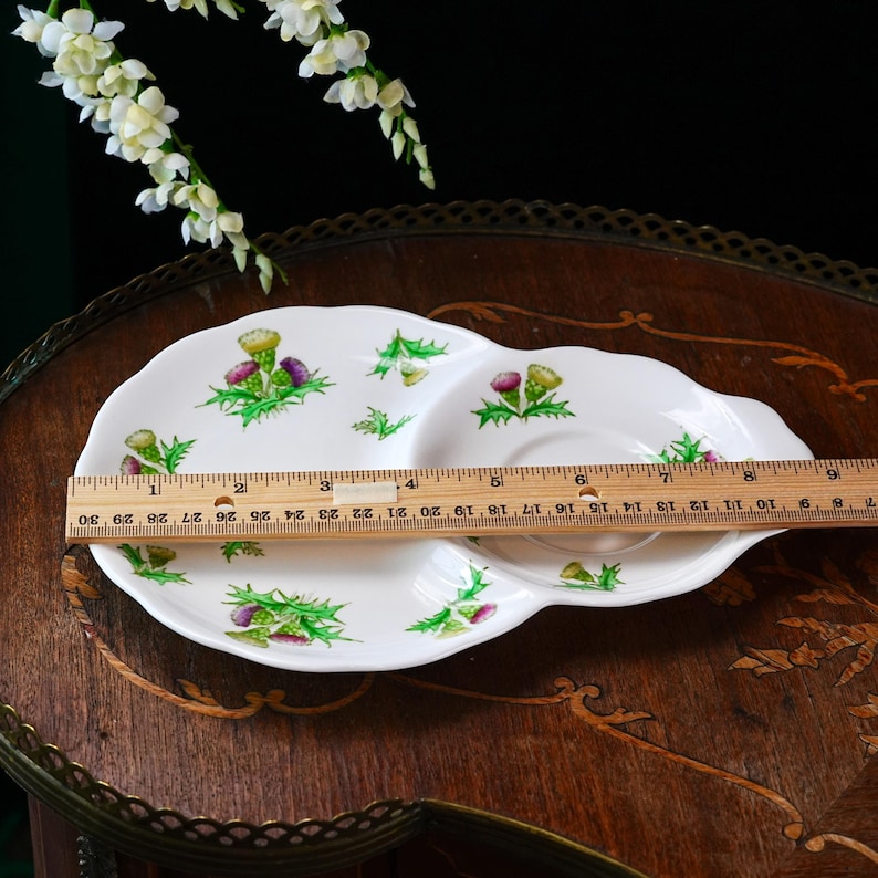 Puede incluir: Plato de cer&aacute;mica blanca con un dise&ntilde;o dividido y un patr&oacute;n de cardo. El plato mide aproximadamente 30 cm de largo. El dise&ntilde;o del cardo presenta acentos morados y verdes. El plato est&aacute; sobre una mesa de madera.