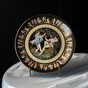 May include: Decorative black plate with a gold rim featuring a classical scene of figures in colorful attire. The central image depicts two figures, one playing a flute, and the other reclining. The plate's border is adorned with additional figures.
