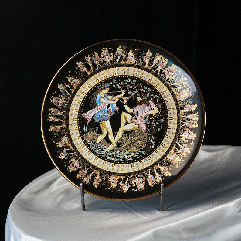 May include: Decorative black plate with a gold rim featuring a classical scene of figures in colorful attire. The central image depicts two figures, one playing a flute, and the other reclining. The plate's border is adorned with additional figures.