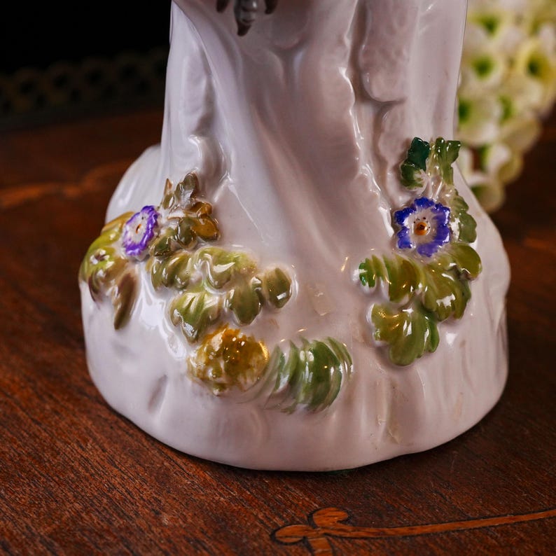 May include: A white porcelain figurine base with hand-painted floral details. The base features green leaves and small blue and purple flowers. The figurine is on a dark wooden surface.