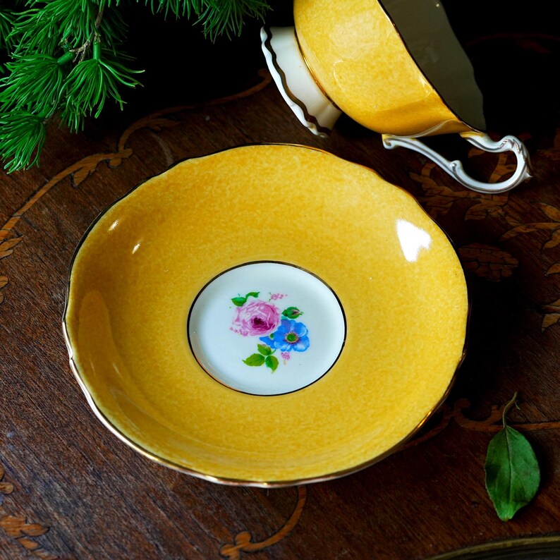 May include: A yellow teacup saucer with a floral design in the centre. The saucer has a gold trim and a textured surface. A matching yellow teacup is in the background. The floral design features pink and blue flowers.