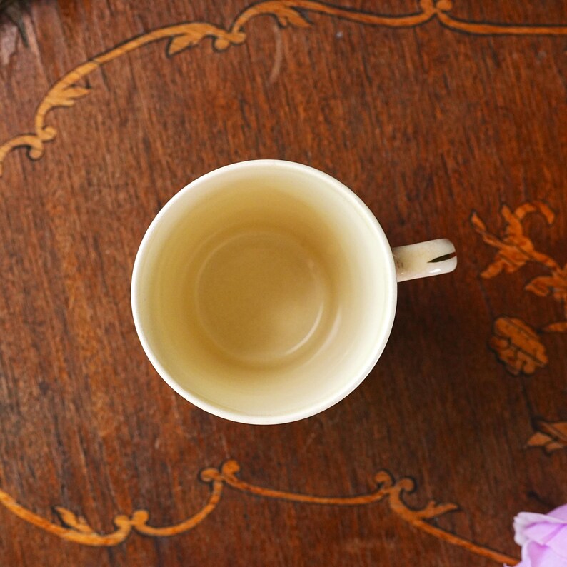Puede incluir: Vista a&eacute;rea de una taza de t&eacute; de cer&aacute;mica blanca rota vac&iacute;a, con un asa peque&ntilde;a. La taza est&aacute; sobre una superficie de madera marr&oacute;n oscuro con patrones incrustados decorativos. El interior de la taza es de color crema claro.