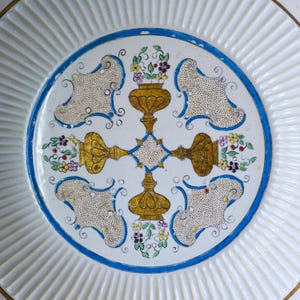 May include: A white ceramic plate with a fluted rim and a central design. The design features a blue border, gold accents, and floral motifs in purple, yellow, and green. The plate has a diameter of approximately 25 cm.