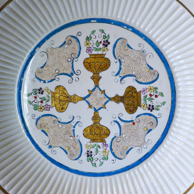 May include: A white ceramic plate with a fluted rim and a central design. The design features a blue border, gold accents, and floral motifs in purple, yellow, and green. The plate has a diameter of approximately 25 cm.