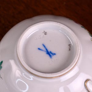 Puede incluir: Base de taza de t&eacute; de porcelana blanca con borde dorado. La parte inferior presenta una marca de espadas cruzadas azules, el n&uacute;mero 207 y otras marcas. La taza tiene un delicado dise&ntilde;o floral.