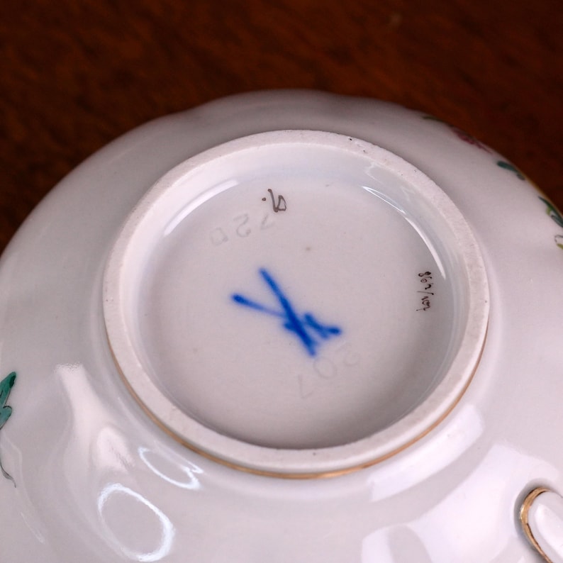 Puede incluir: Base de taza de t&eacute; de porcelana blanca con borde dorado. La parte inferior presenta una marca de espadas cruzadas azules, el n&uacute;mero 207 y otras marcas. La taza tiene un delicado dise&ntilde;o floral.