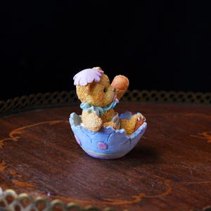 May include: A small, detailed figurine of a teddy bear emerging from a light blue Easter egg. The bear is tan with a pink hat and blue bow. The egg has purple and pink accents. The figurine is on a wooden surface.