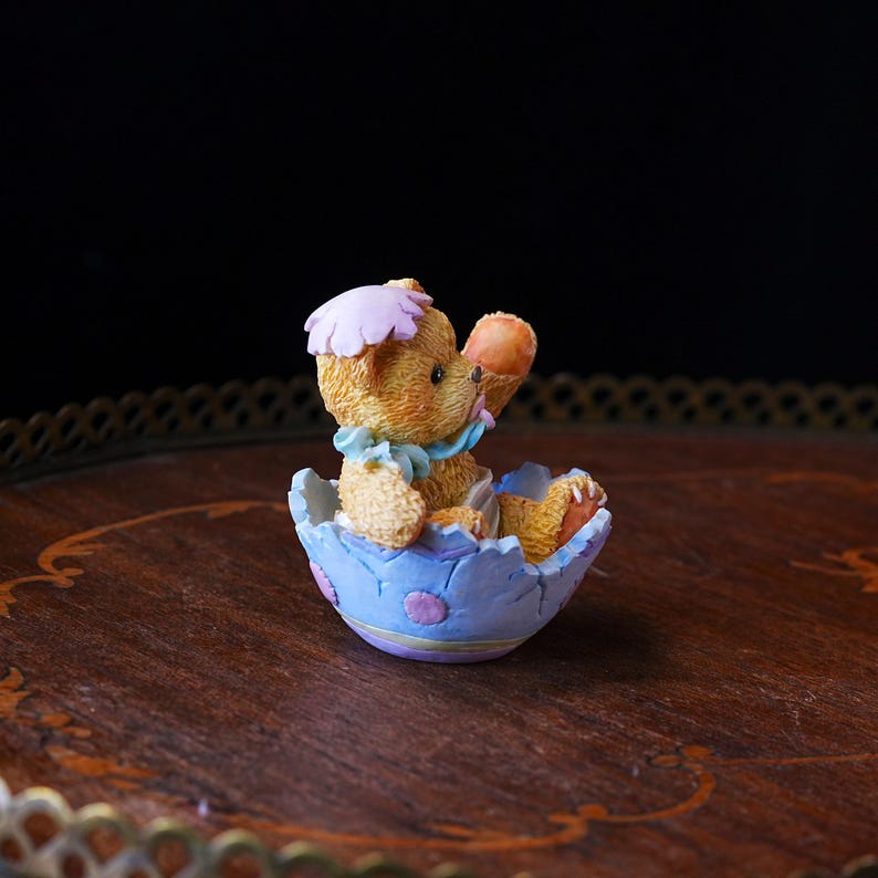 May include: A small, detailed figurine of a teddy bear emerging from a light blue Easter egg. The bear is tan with a pink hat and blue bow. The egg has purple and pink accents. The figurine is on a wooden surface.