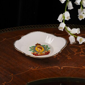 May include: A white porcelain dish with a scalloped edge and gold trim. The center features a colorful royal emblem with a profile portrait of Queen Elizabeth II, surrounded by a green wreath and a red banner with the text DIEU ET MON DROIT.