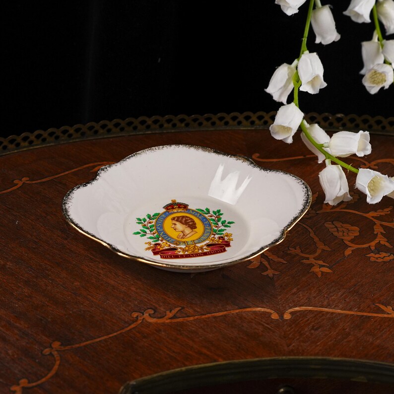 May include: A white porcelain dish with a scalloped edge and gold trim. The center features a colorful royal emblem with a profile portrait of Queen Elizabeth II, surrounded by a green wreath and a red banner with the text DIEU ET MON DROIT.