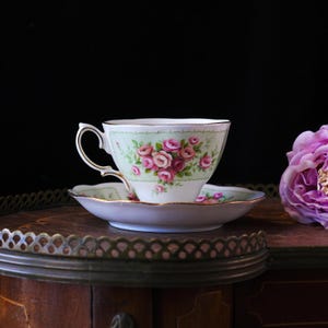 K&ouml;nnte beinhalten: Ein Vintage-Tee-Tassen- und Untertassen-Set. Die Tasse und Untertasse sind wei&szlig; mit einem hellgr&uuml;nen Rand und einem Blumenmuster aus rosa Rosen. Die Tasse hat einen goldenen Henkel und Zierrat. Das Set steht auf einer Holzoberfl&auml;che.