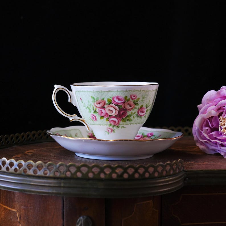 K&ouml;nnte beinhalten: Ein Vintage-Tee-Tassen- und Untertassen-Set. Die Tasse und Untertasse sind wei&szlig; mit einem hellgr&uuml;nen Rand und einem Blumenmuster aus rosa Rosen. Die Tasse hat einen goldenen Henkel und Zierrat. Das Set steht auf einer Holzoberfl&auml;che.