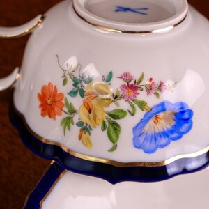 Puede incluir: Taza y platillo de porcelana blanca con dise&ntilde;o floral. La taza presenta un borde azul y ribete dorado, con un colorido arreglo de flores naranjas, amarillas, rosas y azules. El platillo es parcialmente visible.