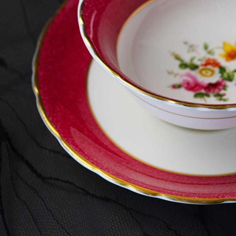 K&ouml;nnte beinhalten: Ein Porzellan-Tassenset mit Untertasse, das einen tief magentafarbenen Rand mit strukturiertem Muster und goldfarbenen, gewellten R&auml;ndern aufweist. Die wei&szlig;e Tasse zeigt in der Mitte ein buntes Blumenbouquet mit rosa, orangen und gelben Bl&uuml;ten.