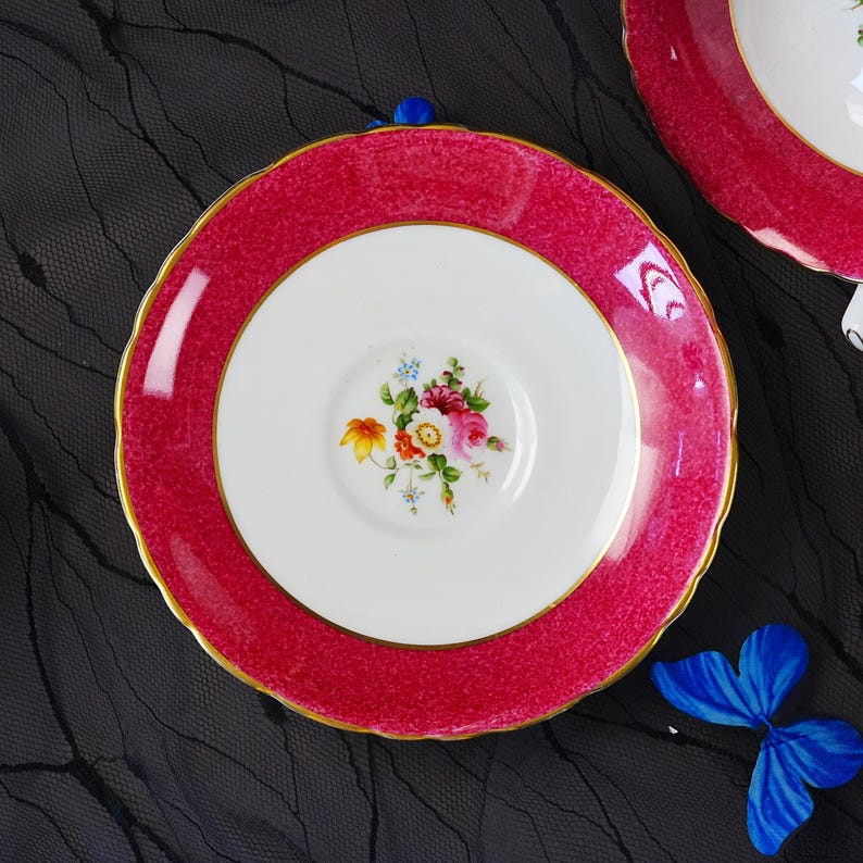 K&ouml;nnte beinhalten: Eine runde Untertasse aus Porzellan mit einem breiten, texturierten magentafarbenen Rand und einem goldfarbenen, gewellten Rand. Die Mitte ist wei&szlig; und mit einem kleinen, bunten Blumenstrau&szlig; aus rosa, gelben und blauen Bl&uuml;ten mit gr&uuml;nen Bl&auml;ttern verziert.