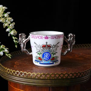 May include: A white ceramic mug with silver lion-shaped handles. The mug is decorated with a crest, a crown, crossed weapons, and the words "SILVER WEDDING". The design includes red and blue details.