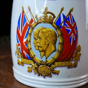 May include: White ceramic mug with a decorative emblem featuring two gold-toned profiles, a crown, and Union Jack flags. The emblem is surrounded by floral accents and the dates 1910 and 1935.