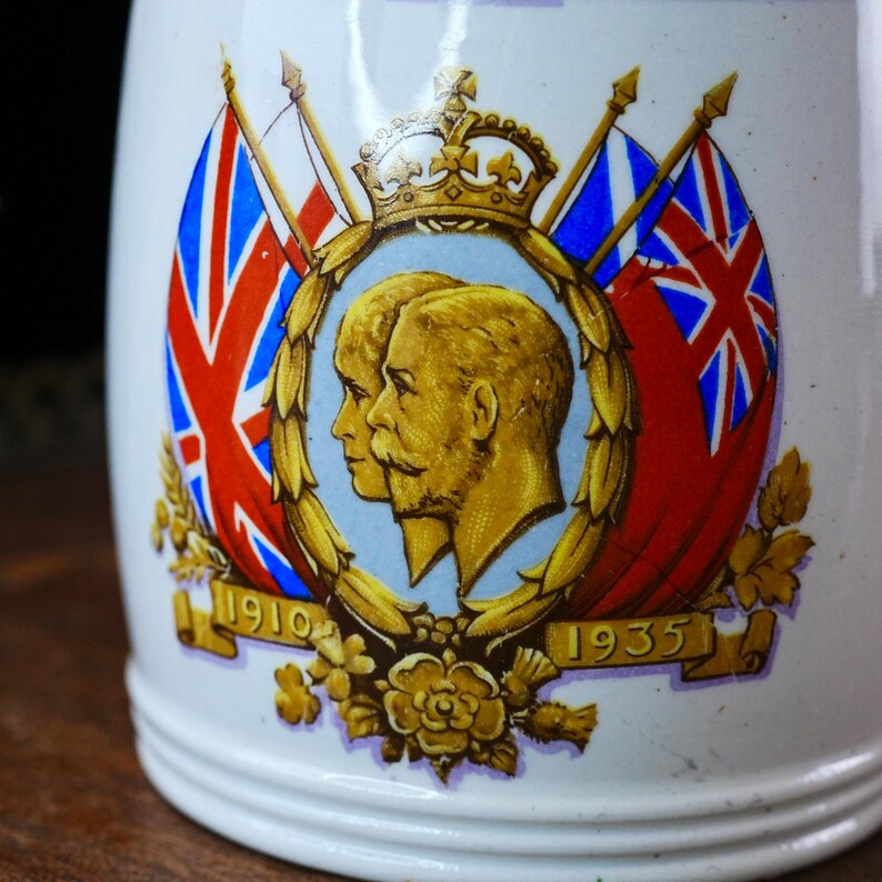 May include: White ceramic mug with a decorative emblem featuring two gold-toned profiles, a crown, and Union Jack flags. The emblem is surrounded by floral accents and the dates 1910 and 1935.