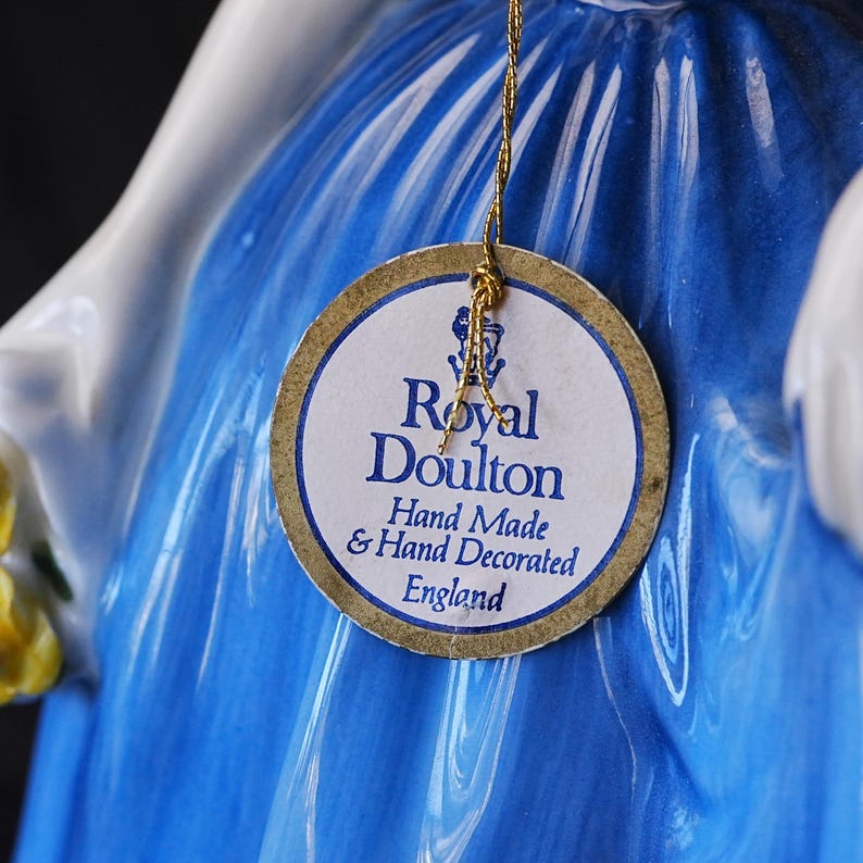 May include: A porcelain figurine wearing a vibrant blue dress with a glossy finish. A round paper tag is attached by a gold string, featuring the text Royal Doulton, Hand Made & Hand Decorated, England, with a small lion and crown logo above.
