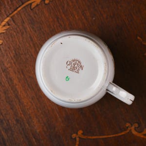 Może przedstawiać: Sp&oacute;d białego ceramicznego kubka z brązowym nadrukiem znaku producenta o treści A.B.J. CRAFTON CHINA MADE IN ENGLAND oraz małą zieloną literą G wybita poniżej. Kubek posiada białe ucho z cienkim złotym wykończeniem na krawędziach.