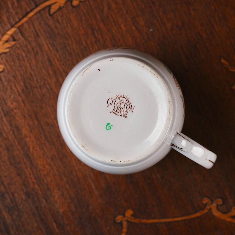 Może przedstawiać: Sp&oacute;d białego ceramicznego kubka z brązowym nadrukiem znaku producenta o treści A.B.J. CRAFTON CHINA MADE IN ENGLAND oraz małą zieloną literą G wybita poniżej. Kubek posiada białe ucho z cienkim złotym wykończeniem na krawędziach.