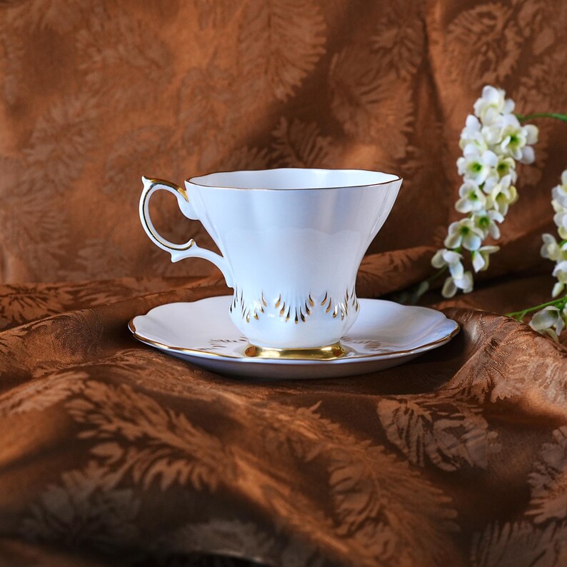 May include: A white teacup and saucer set with gold trim. The teacup has a curved handle and a scalloped rim. The saucer has a matching gold rim and a floral design. The set is displayed on a brown patterned fabric.
