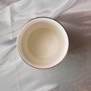 Op de afbeelding: Een cr&egrave;mekleurige, ronde kom met een gouden rand. De kom is leeg en lijkt van keramiek of porselein te zijn. De afbeelding is genomen vanuit een vogelperspectief en toont de binnenkant van de kom.