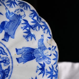 Puede incluir: Una secci&oacute;n de un plato de porcelana blanca con borde festoneado, con ilustraciones azules de figuras y hojas de bamb&uacute;. Las figuras visten ropa tradicional. El plato tiene una muesca en el borde.