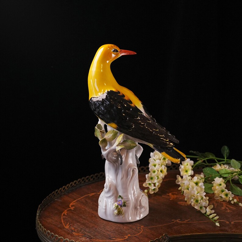 May include: A ceramic bird figurine with a yellow head and chest, black wings, and a red beak. The bird is perched on a white tree trunk with green leaves and small purple flowers. The figurine is on a wooden table.