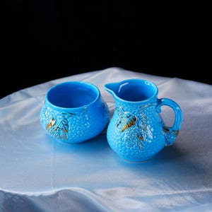 Op de afbeelding: Een felblauwe keramische melkkan en een bijpassende suikerpot. Beide stukken hebben een textuur met een korrelig oppervlak en reli&euml;fmotieven van bladeren en bessen, geaccentueerd met goudkleurige verf. De kan heeft een handvat en een tuit.