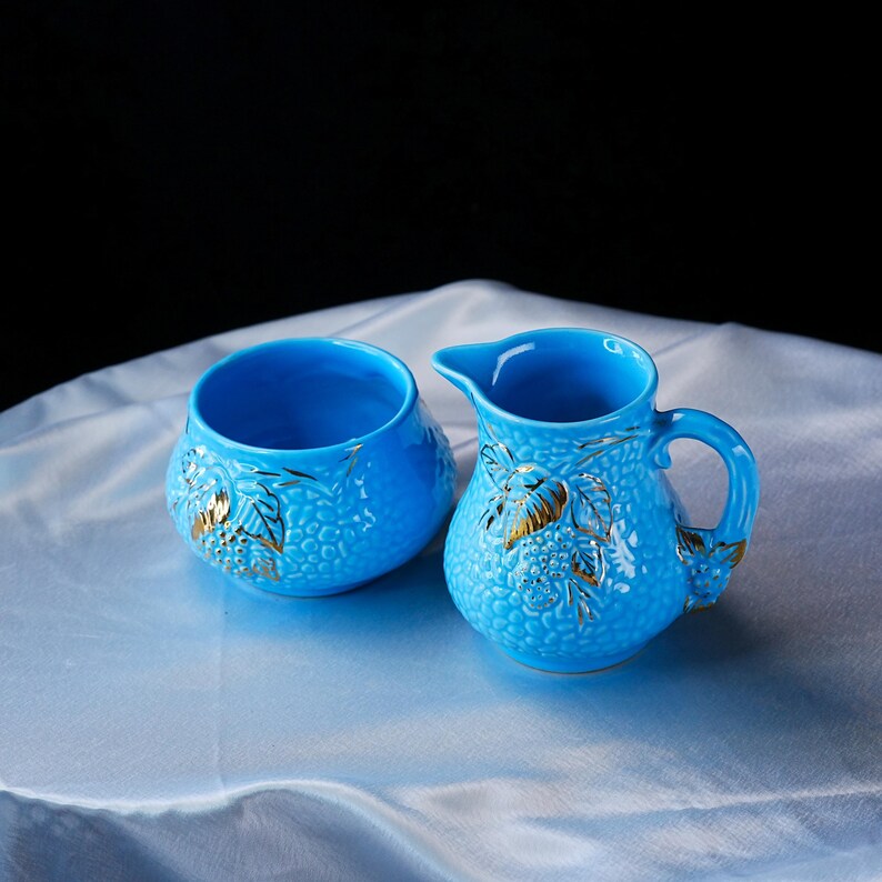 Op de afbeelding: Een felblauwe keramische melkkan en een bijpassende suikerpot. Beide stukken hebben een textuur met een korrelig oppervlak en reli&euml;fmotieven van bladeren en bessen, geaccentueerd met goudkleurige verf. De kan heeft een handvat en een tuit.