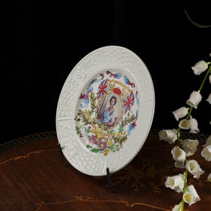 Op de afbeelding: Een witte keramische herdenkingsbord met een centraal portret van koningin Elizabeth II, omringd door kleurrijke bloemillustraties, twee Britse vlaggen en de tekst 25th Anniversary of HM Queen Elizabeth II 1952-1977 met een gestructureerde rand.