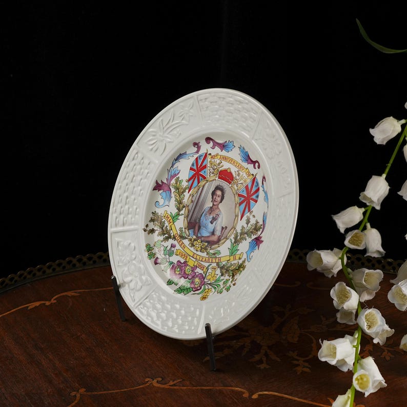 Op de afbeelding: Een witte keramische herdenkingsbord met een centraal portret van koningin Elizabeth II, omringd door kleurrijke bloemillustraties, twee Britse vlaggen en de tekst 25th Anniversary of HM Queen Elizabeth II 1952-1977 met een gestructureerde rand.