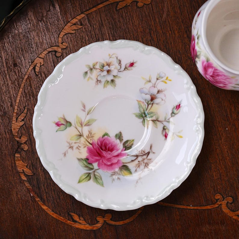 May include: A white porcelain saucer with a scalloped edge, decorated with a pink rose and floral pattern. The design features white and yellow flowers with green leaves. Part of a tea set.