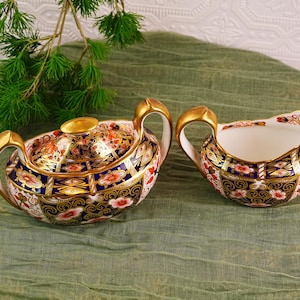 May include: A decorative porcelain tea set featuring a covered sugar bowl and a creamer. The set has a white base with intricate floral and geometric patterns in red, blue, and gold. The handles and lid finial are gold.