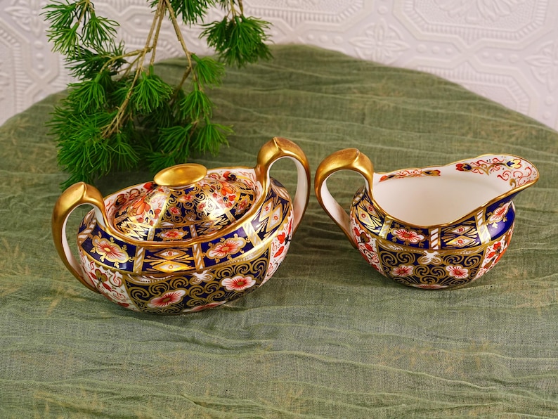 May include: A decorative porcelain tea set featuring a covered sugar bowl and a creamer. The set has a white base with intricate floral and geometric patterns in red, blue, and gold. The handles and lid finial are gold.