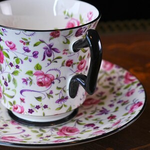 May include: A white teacup and saucer set decorated with pink roses, purple flowers, and green foliage. The teacup features a black handle and rim. The saucer mirrors the floral design with a black edge, perfect for tea time.