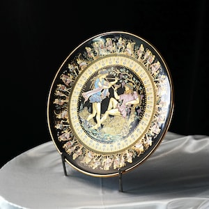 May include: A decorative black plate with a gold rim and a detailed illustration of figures in classical attire. The plate features a central scene with two figures and a border with additional figures. The plate is displayed on a metal stand.