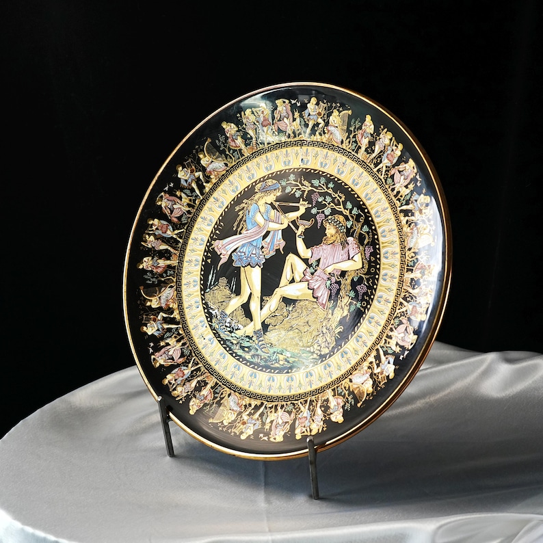 May include: A decorative black plate with a gold rim and a detailed illustration of figures in classical attire. The plate features a central scene with two figures and a border with additional figures. The plate is displayed on a metal stand.