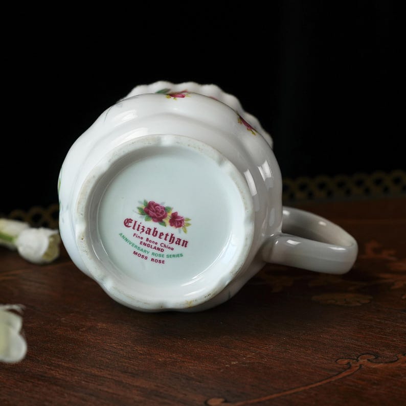Peut inclure: Une tasse en porcelaine fine blanche avec un bord festonn&eacute; et une anse. La base pr&eacute;sente un motif floral et le texte : Elizabethan, Fine Bone China, England, Anniversary Rose Series, Moss Rose. La tasse a une finition lisse et brillante.