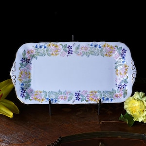 以下が含まれることがあります： ピンク、黄色、青の花のフローラルボーダーが付いた長方形の白い磁器のサービングトレイ。トレイは金色のトリムで、黒いスタンドに展示されています。背景は暗いです。