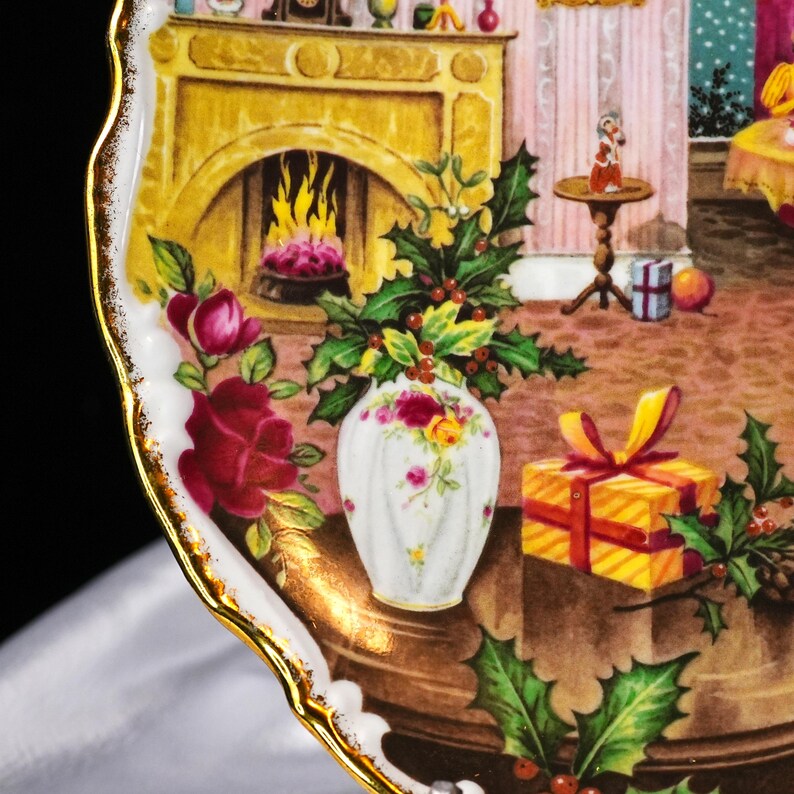 Pu&ograve; includere: Un piatto decorativo con bordo dorato raffigurante una scena d'interni dettagliata. Un camino con fuoco acceso, un vaso di fiori, un regalo avvolto e rametti di agrifoglio sono visibili. Il design del piatto include rose e un tema festivo natalizio.