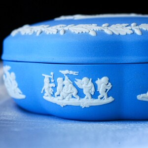 May include: A bright blue ceramic lidded box featuring white relief ornamentation. The lid displays a border of oak leaves and acorns, while the side of the base showcases a detailed frieze of small winged figures in various poses.