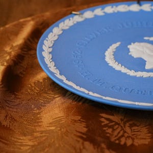 May include: A blue decorative plate with white relief detailing. The plate features a white laurel wreath border and text that reads "E.M. QUEEN 1952-SILVER JUBILEE". A white profile of a person is also visible.